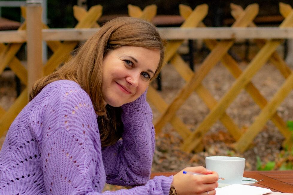 Fine sitzt draußen an einem Holztisch, schaut freundlich in die Kamera. Auf dem Tisch sind zwei Notizhefte und eine Kaffeetasse.