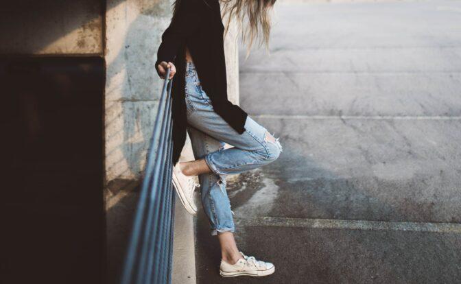Eine Frau in blauer Jeans, einem schwarzen langen Oberteil und weißen Turnschuhen lehnt allein an einem Geländer, den Blick nach unten.