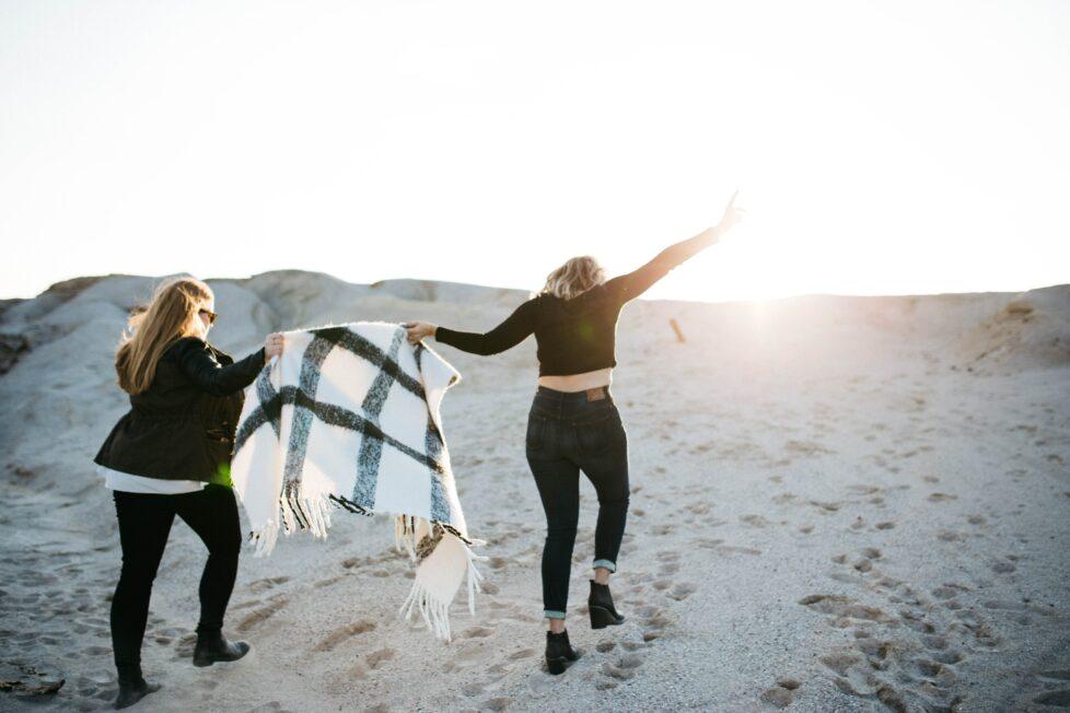 Zwei Frauen gehen auf einer Düne gemeinsam in Richtung Sonne, es wirkt wie ein Neuanfang - einfach anfangen.