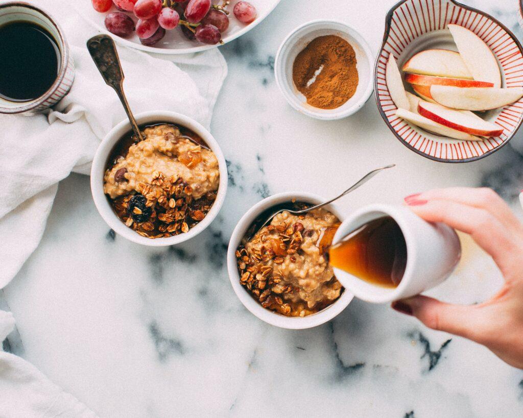Das Bild zeigt zwei kleine Schalen mit Porridge, Weintrauben, Äpfeln und eine Karaffe mit Ahornsirup.