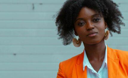 Das Bild zeigt eine junge schwarze Frau in einem orangenem Blazer, die selbstbewusst in die Kamera blickt. Ihre Körperhaltung sagt Nein!