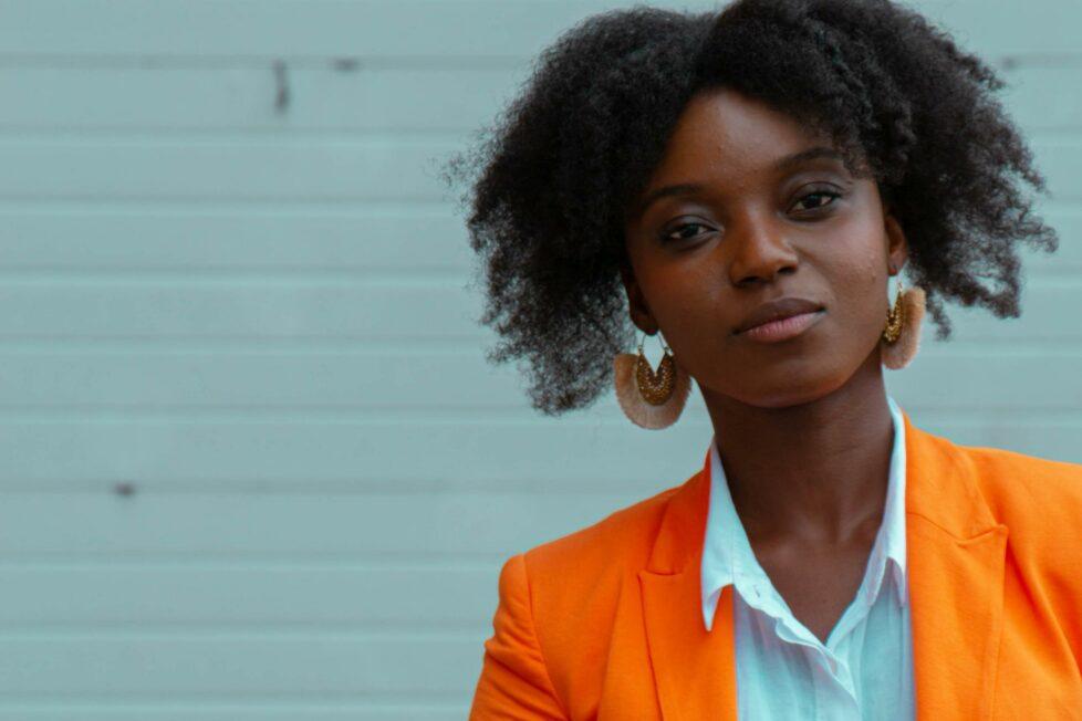 Das Bild zeigt eine junge schwarze Frau in einem orangenem Blazer, die selbstbewusst in die Kamera blickt. Ihre Körperhaltung sagt Nein!