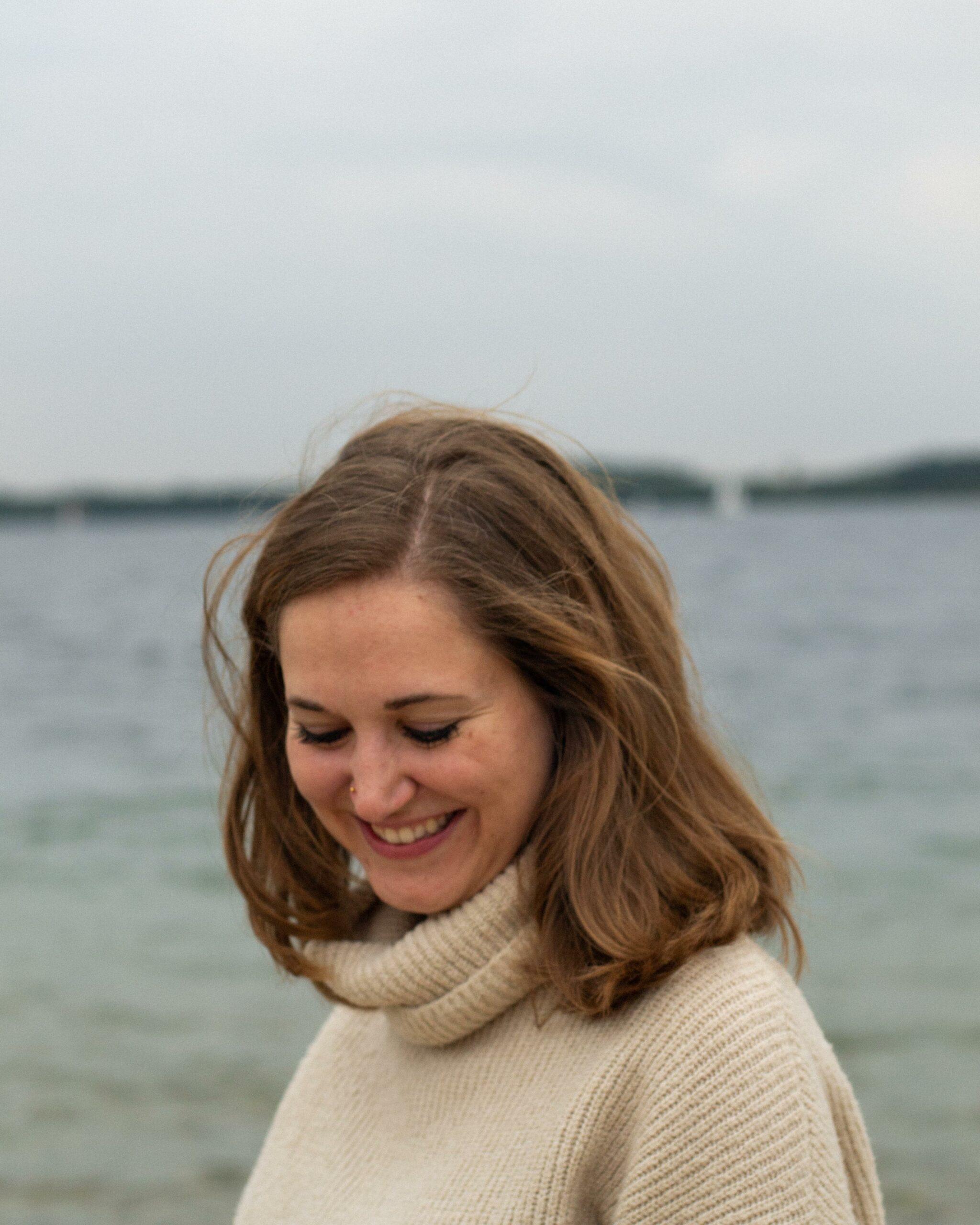 Das Bild zeigt Fine von True Good Things. Die junge Frau trägt einen hellen Pullover und lächelt. Sie steht vor einem See.