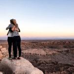 Zwei Freundinnen umarmen sich wertschätzend auf einem Hügel, in Hintergrund ist eine riesige Wüste und die untergehende Sonne.