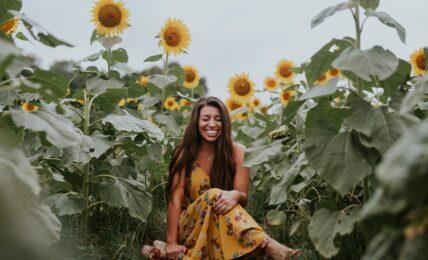 Frau sitzt lachend und erfüllt draußen zwischen hohen Sonnenblumen.