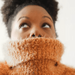 Das Bild zeigt eine schwarze Frau in einem orangenem Rollkragenpullover, den sie bis unter die Nase gezogen hat - aus Angst und Vorfreude zum beruflichen Neuanfang.