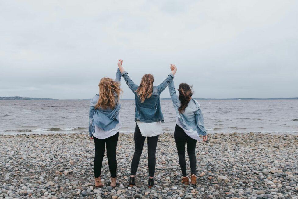Das Bild zeigt drei Frauen am Meer, die vor Freude die Arme in die Luft heben und das Ankommen und Weitermachen von True Good Things feiern.