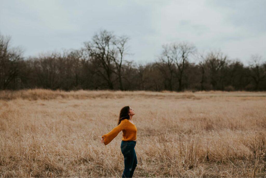 Das Bild zeigt eine junge Frau mit braunem Haar und orangefarbenen Pullover, die in einem Kornfeld steht und ihren Körper in den Gegenwind hält, um zu zeigen, dass sie Distanz zur Arbeit bewahrt.