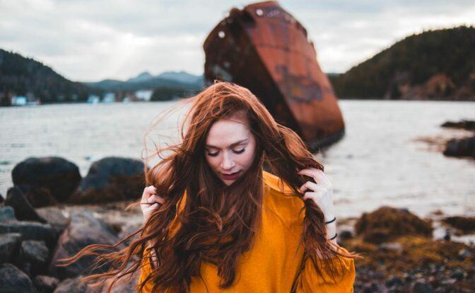 Das Bild zeigt eine Frau mit langen roten Haaren und einem senffarbenen Pullover am Strand, die sich Gedanken darüber macht, was wirklich wichtig ist im Leben.