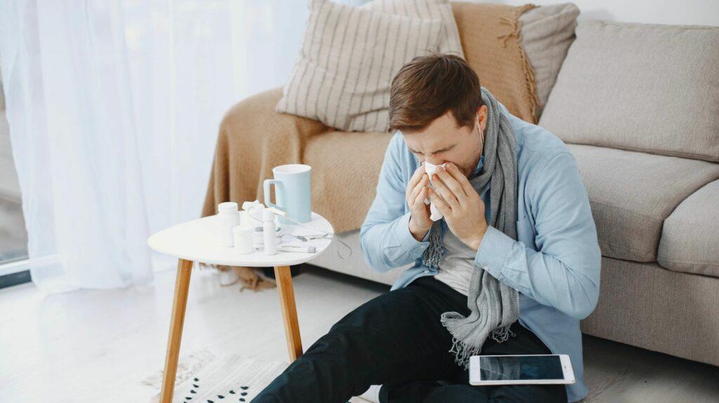 Das Bild zeigt einen Mann mit kurzen braunen Haaren vor seinem Sofa sitzend, der sich die Nase putzt und sich überlegt, ob er so krank zur Arbeit gehen soll.