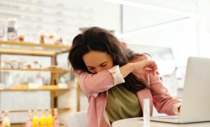 Das Bild zeigt eine Frau mit braunen Haaren am Arbeitsplatz vor ihrem Laptop, die in ihren Ellenbogen niest, weil sie krank zur Arbeit gekommen ist.