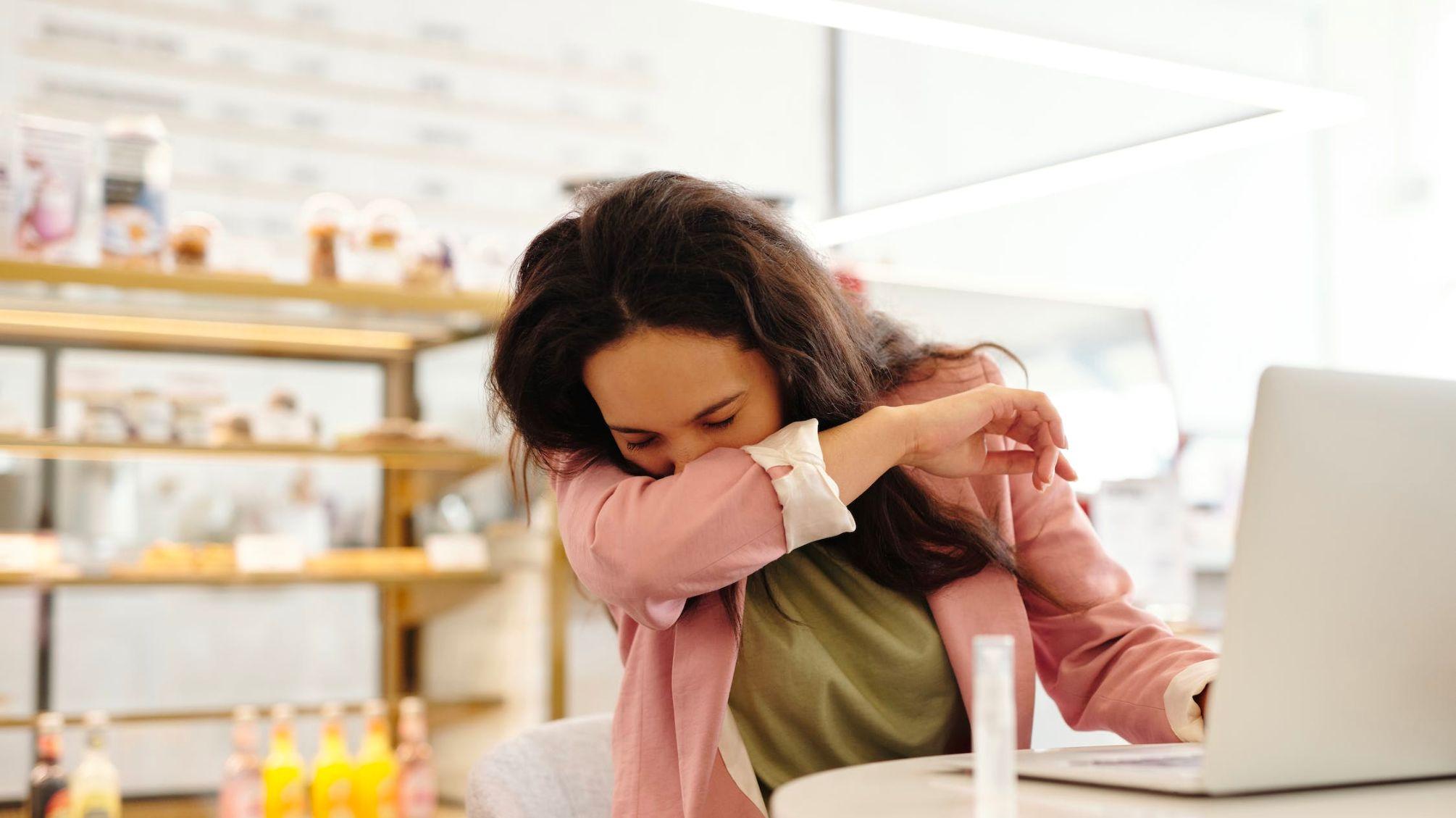 Das Bild zeigt eine Frau mit braunen Haaren am Arbeitsplatz vor ihrem Laptop, die in ihren Ellenbogen niest, weil sie krank zur Arbeit gekommen ist.