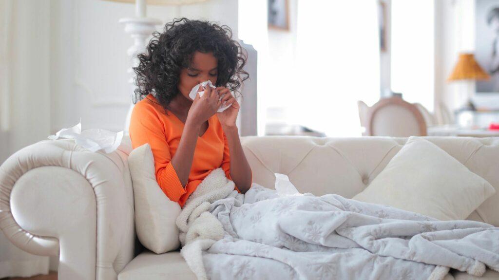 Das Bild zeigt eine schwarze Frau, die auf ihrem Sofa sitzt und sich die Nase putzt und dabei überlegt, ob Kranksein im Büro in Ordnung ist.